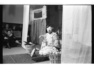 A man is seated on a tarp runway and he is blindfolded. He is surrounded by several small objects and two baskets on either side. Curtains drape down from the ceiling. There is a curtain with a frame that rests on top of it behind the man. The background features a seated audience. The image is in black and white.