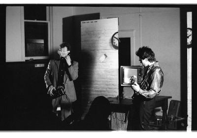 Mike Haslam sings into a touch-tone telephone receiver while fingering the keypad held at his waist. He wears a suit and sunglasses. He is joined onstage by someone playing bass guitar. They perform in front of a television monitor and one-half of the freestanding brick wall backdrop. 