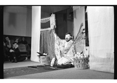 A man is seated on a tarp runway and he is blindfolded. He is surrounded by several small objects and two baskets on either side of him, one with fruits in it. The man holds something in his right hand and has his arm raised. Curtains drape down from the ceiling. There is a curtain with a frame that rests on top of it, adjacent to the man. The background features an audience. The image is in black and white.