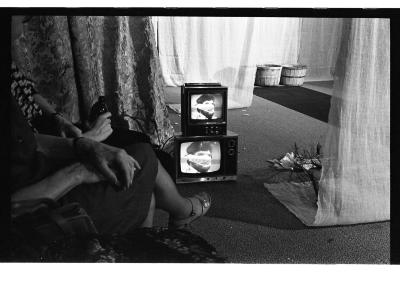 Someone sits with their legs crossed in front of a performance set that includes two stacked CRT televisions that display the same image of a man with a lace muzzle, draped curtains, a tarp runway, and two baskets.