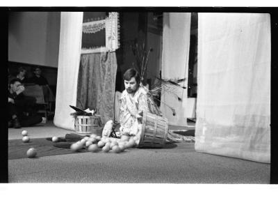 A man is seated on a tarp runway. He is surrounded by several small objects and two baskets on either side of him, one of which has fruit spilling out of it. Curtains drape down from the ceiling. There is a curtain with a frame that rests on top of it, adjacent to the man. The background features an audience. The image is in black and white.