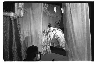 A performer wearing a kimono-like robe is framed by sheer curtains and a banner hung from the ceiling.. They stand with their back to the camera and their long sleeves dramatically billow behind them. The foreground features the back of a man’s head.