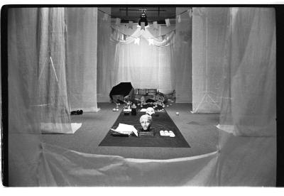 Various objects are arranged on a tarp runway. framed by translucent curtains draping down from the ceiling. Banners and lights loom over this set. 