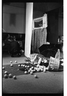 There are several objects scattered on a tarp runway, including a basket of spilled fruit, a Noh mask, and an open umbrella. Curtains drape down from the ceiling. There is a curtain with a frame that rests on top of it. The audience shifts their gaze to this curtain with a frame on top of it.