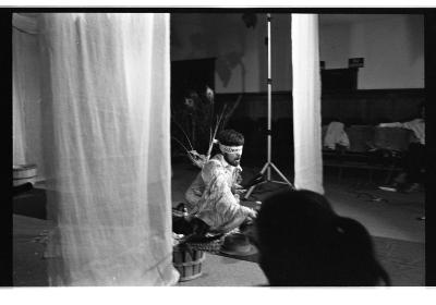 A man is seated on a tarp runway and he is blindfolded. He wears a kimono-like robe with peacock feathers extending from the back of the garment. He is surrounded by several small objects and his right arm is extended out in front of them. Curtains drape down from the ceiling. The foreground features a silhouette of an audience-members head. The image is in black and white.