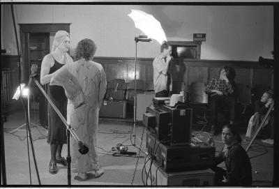 Backstage of a set, arranged with several pieces of camera equipment and a camera crew. The image is in black and white.