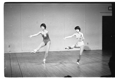 Two women synchronously dance in a studio. Each dancer points their toe out and extends their arms out in front of themselves. They both have short hair, wear leotards, and leg warmers. 