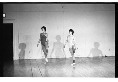 Two women do a pirouette in a studio. They both have short hair, wear leotards, and leg warmers. 