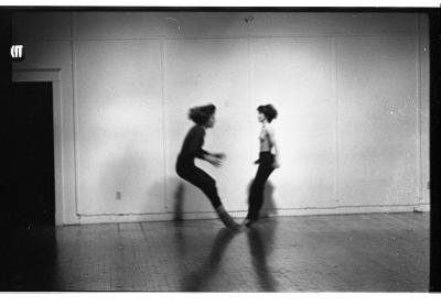 A blurred image of two dancers performing in a studio. They stand two-to-two as they bend and tip backwards.