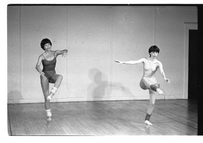 Two women dance in a studio. They have short hair, wear leotards, and leg warmers. The dancer on the left has her knee bent in the air, and the dancer on the right has her leg stretched out in front in air. Both dancers create gestural movements with their arms in the air.