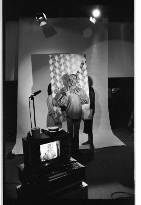 A front-shot of two people hoisting a harlequin backdrop behind a man in a puffed long sleeve shirt, with his open hands raised. His face is painted in a harlequin pattern that matches the backdrop. Behind them is a long blank backdrop that drapes to the floor and two spotlights above. A monitor with a microphone is in the foreground framing the man with the painted face.
The image is in black and white.