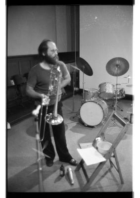 A black-and-white photo of Gunter Christmann holding his trombone. A drum kit is set up behind him, and a wooden folding chair is open in front of him, where a piece of paper and a bowl are positioned. A small bottle of beer and a cylindrical metal object are on the floor near his feet. He wears glasses and is dressed in a T-shirt, long dark pants, and leather shoes. 
