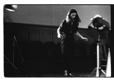 A long-shot of a woman standing as she gazes downward. Her arms gesture outward as if she is attempting to balance herself. The wall in the background is finished with wainscoting in front of it are retractable seats. The foreground depicts blurred images of stage lights.