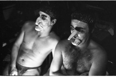 A black-and-white portrait of Martin von Haselberg and Brian Routh. They are both shirtless, and are wearing shower caps and prosthetic noses and chins. Their faces are painted white with black dots to create the effect of stubble. 