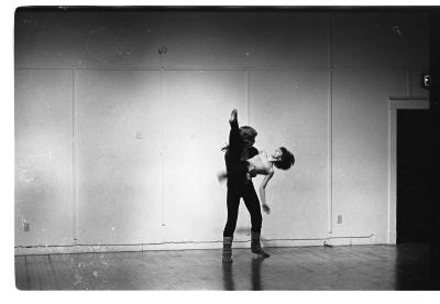 An image of two dancers performing in a studio. One of the dancers lifts the other horizontally against his torso as her leg extends above his own head.