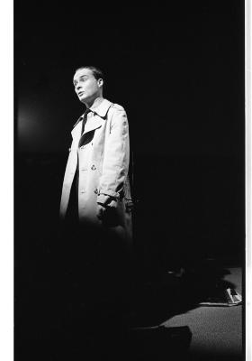 A long-shot of an actor cast in stark lighting. He is wearing a coat and a surprised expression. The image is in black and white.