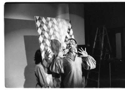 A three-quarter front shot of a person hoisting a harlequin backdrop behind a man in a puffed long sleeve shirt, with his open hands raised. His face is painted in a harlequin pattern that matches the backdrop. The unlit area in the background features more camera equipment and a tall ladder.