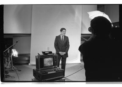 A studio set up featuring photography equipment and a monitor. A man in a suit and tie stands in front of a backdrop, while the photographer in the foreground captures photos of him. The image is in black and white.