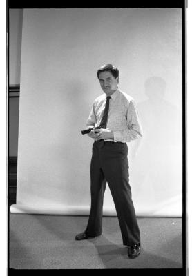 A long-shot featuring a three-quarter profile of a man standing with his legs spread. He's wearing a button up shirt, pants, and shiny shoes. The prop gun he holds against his torso points out. A blank backdrop is draped behind him.