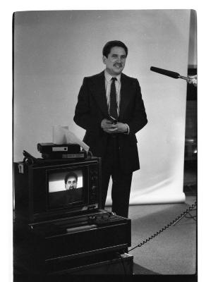 A three-quarter shot featuring a front-profile of a man in a blazer, tie, shiny shoes and pants. The prop gun he holds against his torso points out. A monitor is in front of him and frames a close up of his face, and a blank backdrop is draped behind him. There is a microphone that points from the right-side of the image. The image is in black and white.