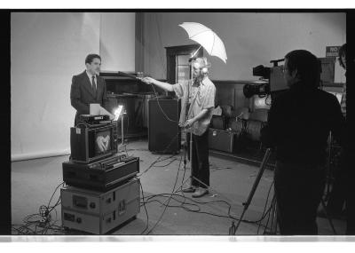 A three-quarter angle-shot of someone pointing a microphone towards an actor, as the moment is captured by the camera-person. The monitor in front of the actor displays his hands together, gesturing down. The background features chairs and other set equipment. The image is in black and white.