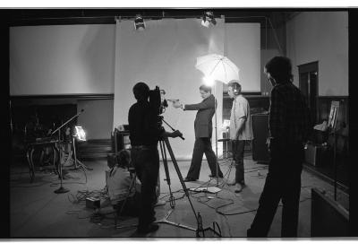 A behind the scenes shot of a camera crew capturing an actor pointing a prop gun to his right. 
Behind them is a long blank backdrop that drapes to the floor and two spotlights above. The image is in black and white.
