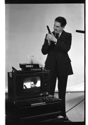 A long-shot featuring a front-profile of a man in a blazer, tie, shiny shoes and pants. He points a prop gun up in the air and gazes toward it. A monitor is in front of him and frames a close up of his face and the prop gun. A blank backdrop is draped behind him. A microphone points from the right-side of the image. The image is in black and white.