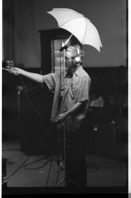 A long-shot of a man wearing headphones and pointing a microphone to his right. A shoot-through umbrella stands in front of him. The image is in black and white.