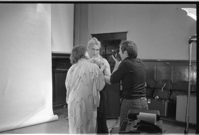 Three people congregate amongst each other, one of them holds a camera directed towards the other two. The background features a room with a long blank backdrop draping to the floor and camera equipment. The image is in black and white.