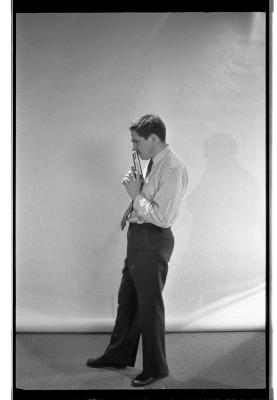 A long-shot featuring a side profile of a man in a shirt, tie, shiny shoes, and pants. He points a prop gun up against his lips. One of his legs extends out in front of the other. A blank backdrop is draped behind him. The image is in black and white.