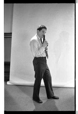 A long-shot featuring a side-profile of a man in a shirt, tie, shiny shoes and pants. He points a prop gun up against his lips, but his head is turned towards the camera taking his picture. One of his legs extends out in front of the other. A blank backdrop is draped behind him. The image is in black and white.