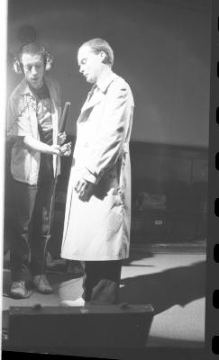 An overexposed shot of a man in headphones pointing a microphone towards an actor as he looks down towards it. The image is in black and white.