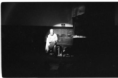 A long shot of a man seated at a grand piano, facing away from the keys. The surrounding area of the room is dark, but he and part of the piano are lit by a bright spotlight.