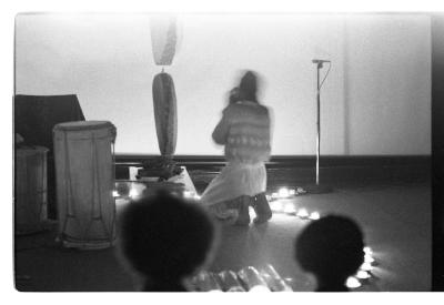 A person with their back to the camera kneels on the floor, blurred as they energetically move. 
They are surrounded by a ring of lit tea lights, and a pair of hide drums hang down from the ceiling. The back of two audience members' heads can be seen in the foreground.
