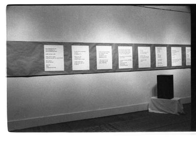 Eight posters with text are mounted in a row on a wall on top of a long strip of dark paper. A speaker is positioned on a plinth draped in fabric on the floor in front.