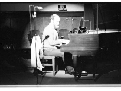 A long shot of a man playing the grand piano. The surrounding area of the room is dark, but he and part of the piano are lit by a bright spotlight. There is a microphone on the stand in the foreground and a no smoking sign in the background.