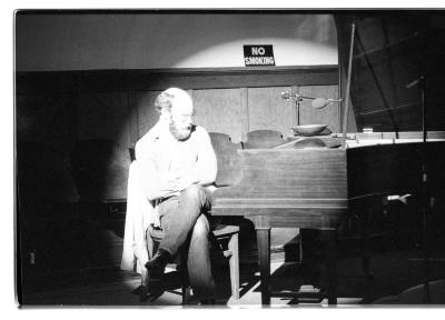 A man is seated at a grand piano with gaze fixed towards the piano keys. The surrounding area of the room is dark, but he and part of the piano are lit by a bright spotlight.