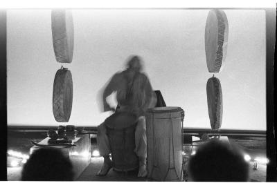 A person is seated and plays a hand drum. Their figure is blurred and they are surrounded by several instruments. They are seated between two rows of drums hanging parallel to one another, each row having two drums. Encompassing them and the instruments is a ring of small lit candles. The foreground depicts the silhouette of two audience members' heads.