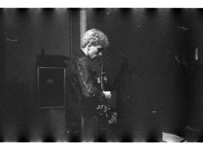 A black and white image with a white border. This grainy photo shows a person playing the guitar with a mass of musical equipment behind them. 