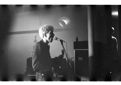 A black and white image.  A person playing guitar and singing into a microphone has blonde hair and is wearing a denim jacket and tight denim pants. 