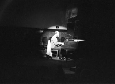 A man seen in profile plays a grand piano. The surrounding area of the room is dark, but he is lit by a bright spotlight.