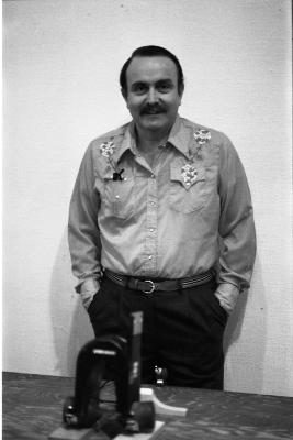 Smiling, Alvin Lucier wears a cowboy shirt and stands with his hands in his pockets. He is behind a wood table that holds a musical device. 