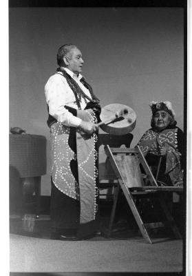 A male Kwakiutl elder dressed in traditional regalia stands in profile and plays a drum. A female elder in regalia is seated behind him. 