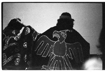 Two performers in traditional Kwakiutl regalia stand with their backs to the camera. One of the performers wears a button blanket with a thunderbird motif.