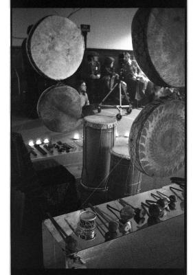 Four round hide drums hang from the ceiling, encompassing other hand drums, mallets, and percussive instruments arranged on the floor and on mirrored tables. This arrangement is surrounded by a ring of lit candles on the floor. Audience observes in the background.