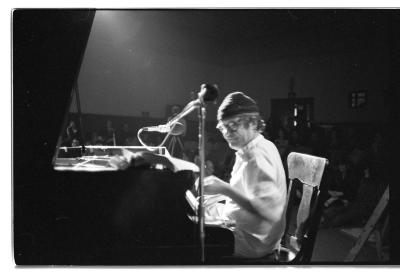 Featured in a side profile, a man is  seated on a wooden chair playing the grand piano. He wears a light coloured tunic and pants, round glasses, and a toque. An audience observes in the background. This image is in black and white.