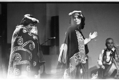 One older woman and one younger woman are dressed in traditional Kwakiutl regalia. The youth looks back with her left palm extending out facing up. The elder behind her has her eyes closed as she claps. In the back there is an older man playing the drum.