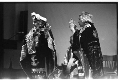 Two elders wearing traditional Kwakiutl regalia perform a dance. They both have their hands open and raised by their faces.