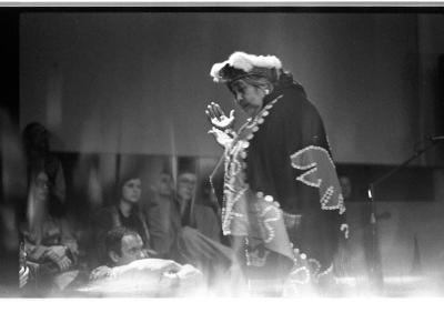 A female Kwakiutl elder dressed in traditional regalia performs a dance. Her gaze shifts down as she claps her hands. In the background a blurred audience watches her performing.
