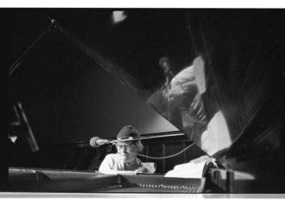 A man wearing a white shirt, a toque, and round glasses plays grand piano. His reflection is mirrored in the glossy piano lid. This image is in black and white.